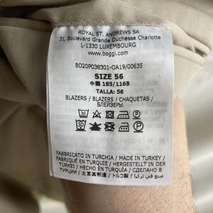 May include: Close-up of a tag from a beige blazer. The tag includes the brand name, address, and size information. The size is listed as 56. Washing instructions are also visible.