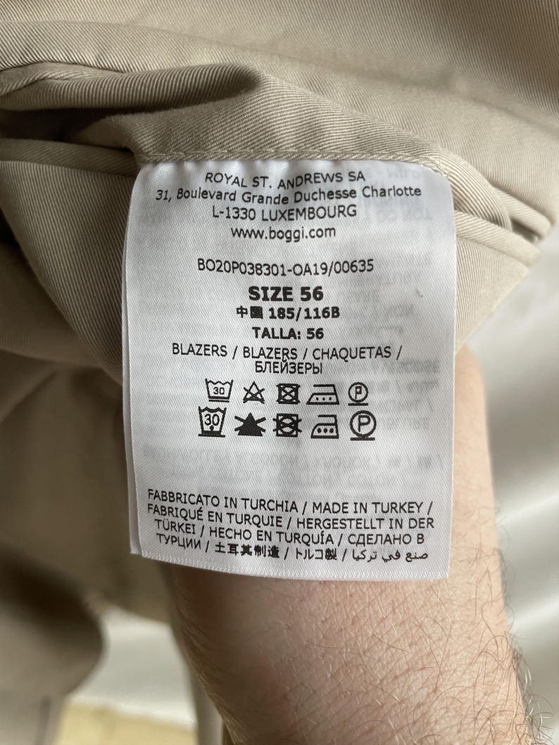 May include: Close-up of a tag from a beige blazer. The tag includes the brand name, address, and size information. The size is listed as 56. Washing instructions are also visible.