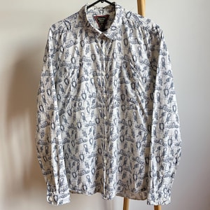 Puede incluir: Una camisa blanca de manga larga con botones y un patrón de nudos marineros azul marino. La camisa tiene cuello y un solo bolsillo en el pecho.