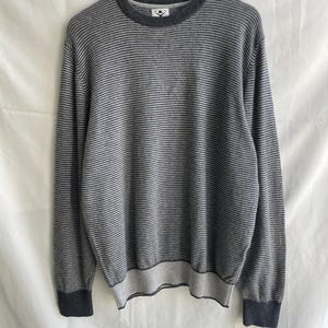 Puede incluir: Un suéter a rayas grises y azul marino con cuello redondo y mangas largas. El suéter tiene dobladillo y puños de canalé. El suéter está colgado en una percha de madera sobre un fondo blanco.