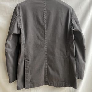 May include: A gray blazer with a notched lapel, two front pockets, and a single-breasted closure. The back of the jacket features a center vent and a tailored fit. The jacket is made of a solid-colored fabric.