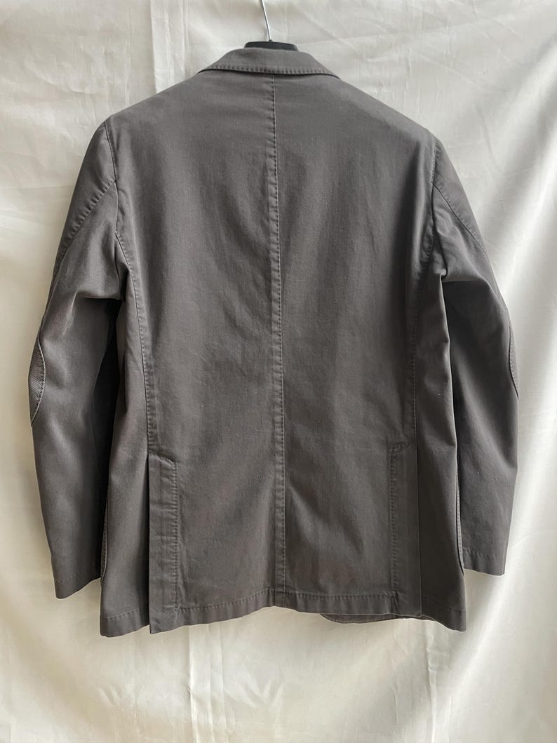 May include: A gray blazer with a notched lapel, two front pockets, and a single-breasted closure. The back of the jacket features a center vent and a tailored fit. The jacket is made of a solid-colored fabric.