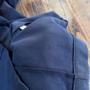 May include: A dark navy blue garment featuring a soft, brushed fleece interior lining. The exterior fabric has a smooth, matte finish with visible reinforced stitching along the seams. A small, square, dark navy patch with a simple face icon is visible.