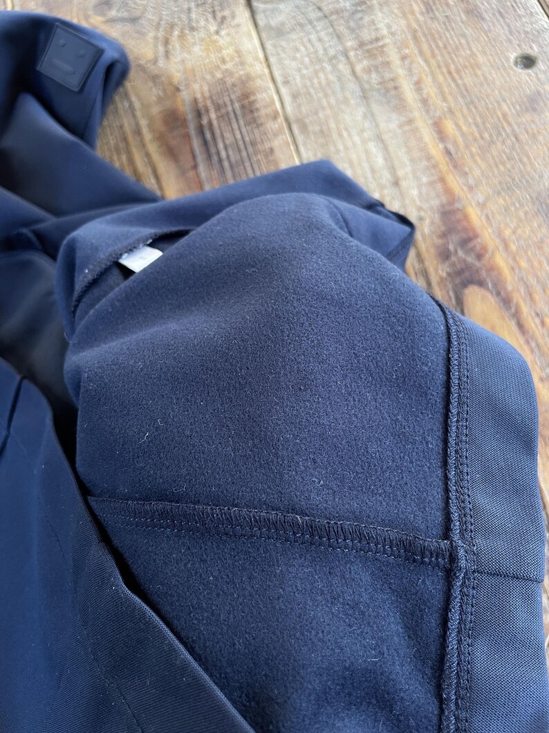 May include: A dark navy blue garment featuring a soft, brushed fleece interior lining. The exterior fabric has a smooth, matte finish with visible reinforced stitching along the seams. A small, square, dark navy patch with a simple face icon is visible.
