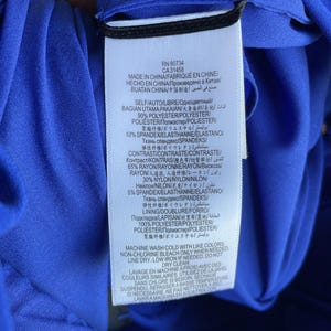 Puede incluir: Etiqueta de una prenda azul con texto en varios idiomas. La etiqueta incluye informaci&oacute;n sobre la composici&oacute;n de la prenda, instrucciones de cuidado y origen. La tela es 90% poli&eacute;ster y 10% spandex.