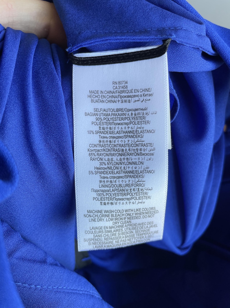 Puede incluir: Etiqueta de una prenda azul con texto en varios idiomas. La etiqueta incluye informaci&oacute;n sobre la composici&oacute;n de la prenda, instrucciones de cuidado y origen. La tela es 90% poli&eacute;ster y 10% spandex.