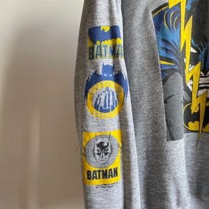 May include: Grey sweatshirt with a large graphic print of Batman on the front. The sleeves have smaller Batman logos in yellow and blue.