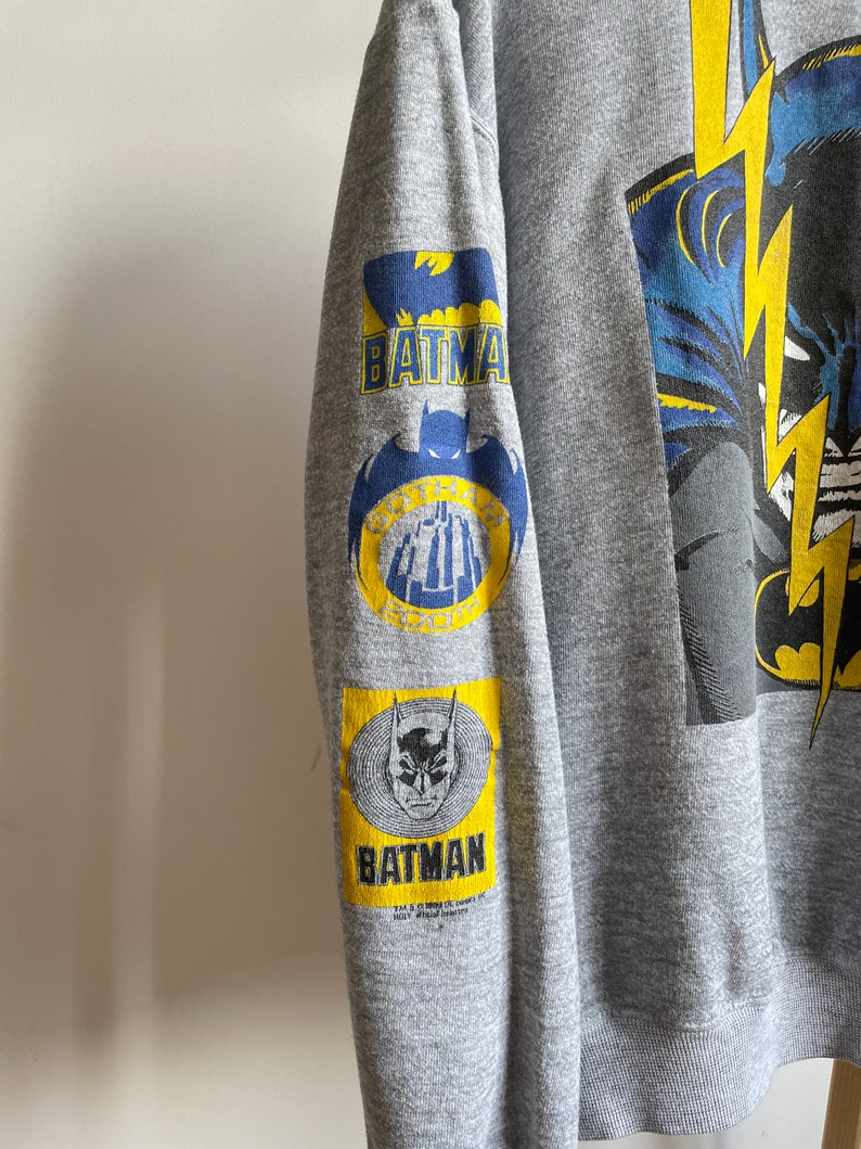 May include: Grey sweatshirt with a large graphic print of Batman on the front. The sleeves have smaller Batman logos in yellow and blue.