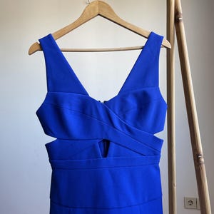 Puede incluir: Un vestido azul vibrante con escote en V y detalles recortados. El vestido est&aacute; colgado en una percha de madera, mostrando su dise&ntilde;o. El vestido tiene un corpi&ntilde;o ajustado y una falda plisada, creando una silueta favorecedora. La tela parece suave y tiene un ligero brillo.