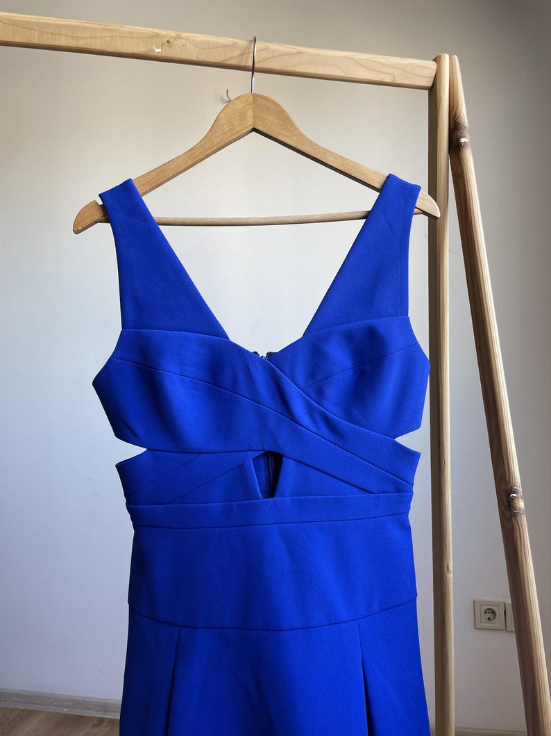 Puede incluir: Un vestido azul vibrante con escote en V y detalles recortados. El vestido est&aacute; colgado en una percha de madera, mostrando su dise&ntilde;o. El vestido tiene un corpi&ntilde;o ajustado y una falda plisada, creando una silueta favorecedora. La tela parece suave y tiene un ligero brillo.