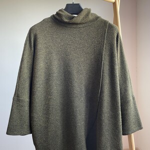 Peut inclure: Haut en maille vert olive avec un col roul&eacute; et des manches trois-quarts. Le haut a un aspect textur&eacute; et une coupe ample. Il est suspendu &agrave; un portant en bois.