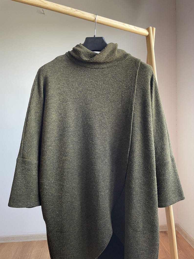 Peut inclure: Haut en maille vert olive avec un col roul&eacute; et des manches trois-quarts. Le haut a un aspect textur&eacute; et une coupe ample. Il est suspendu &agrave; un portant en bois.