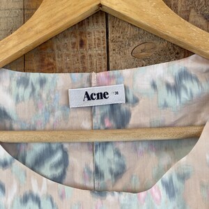 Puede incluir: Una prenda de color beige claro con un patr&oacute;n floral difuminado en tonos azul, verde y rosa. En el escote hay una etiqueta rectangular blanca cosida con el texto negro "Acne" y el n&uacute;mero "38" en un tama&ntilde;o de fuente menor a la derecha.