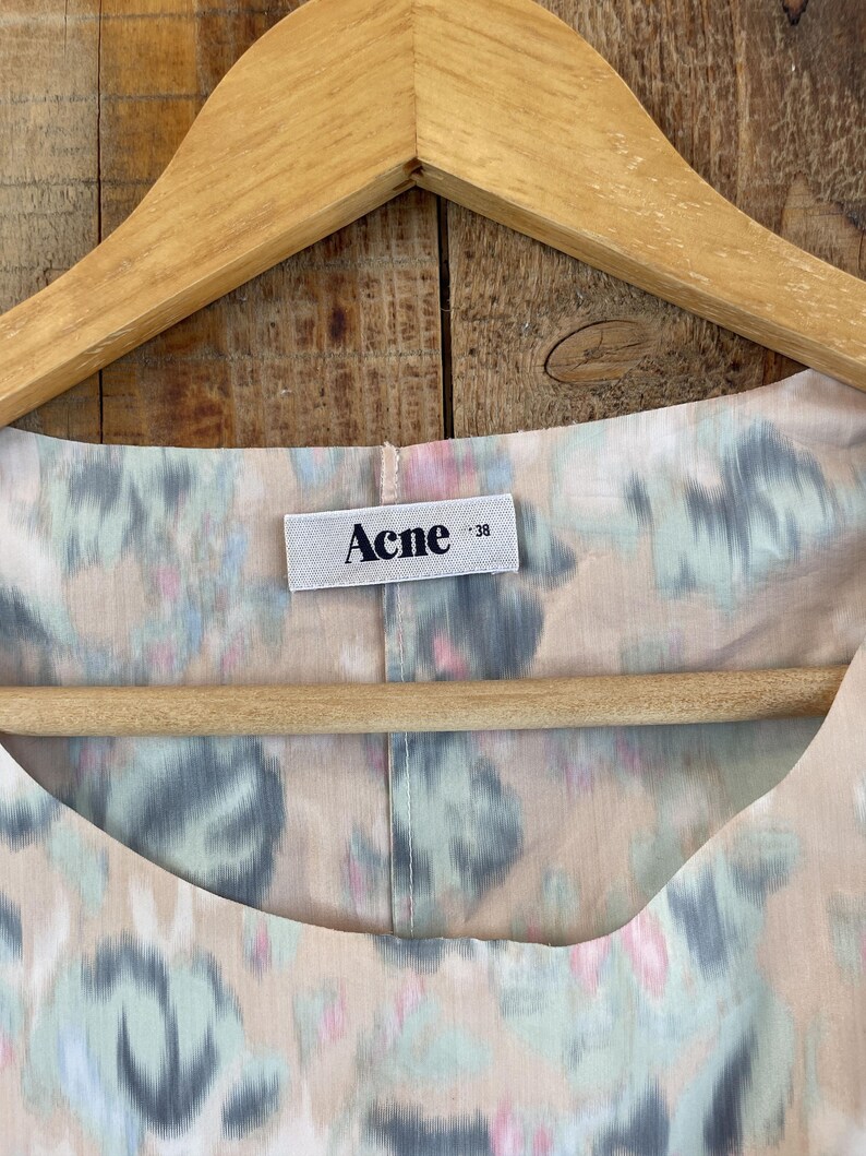 Puede incluir: Una prenda de color beige claro con un patr&oacute;n floral difuminado en tonos azul, verde y rosa. En el escote hay una etiqueta rectangular blanca cosida con el texto negro "Acne" y el n&uacute;mero "38" en un tama&ntilde;o de fuente menor a la derecha.