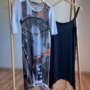 May include: Two dresses hanging on a wooden rack. The dress on the left is white with a black and orange geometric pattern and a sheer overlay. The dress on the right is black and sleeveless.