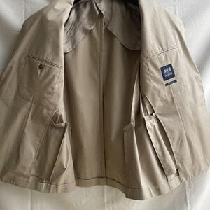 May include: A tan-colored jacket, displayed open, revealing the interior lining and pockets. The jacket is hanging on a black hanger. The interior features a satin-like lining. A label with a logo is visible on the inside.