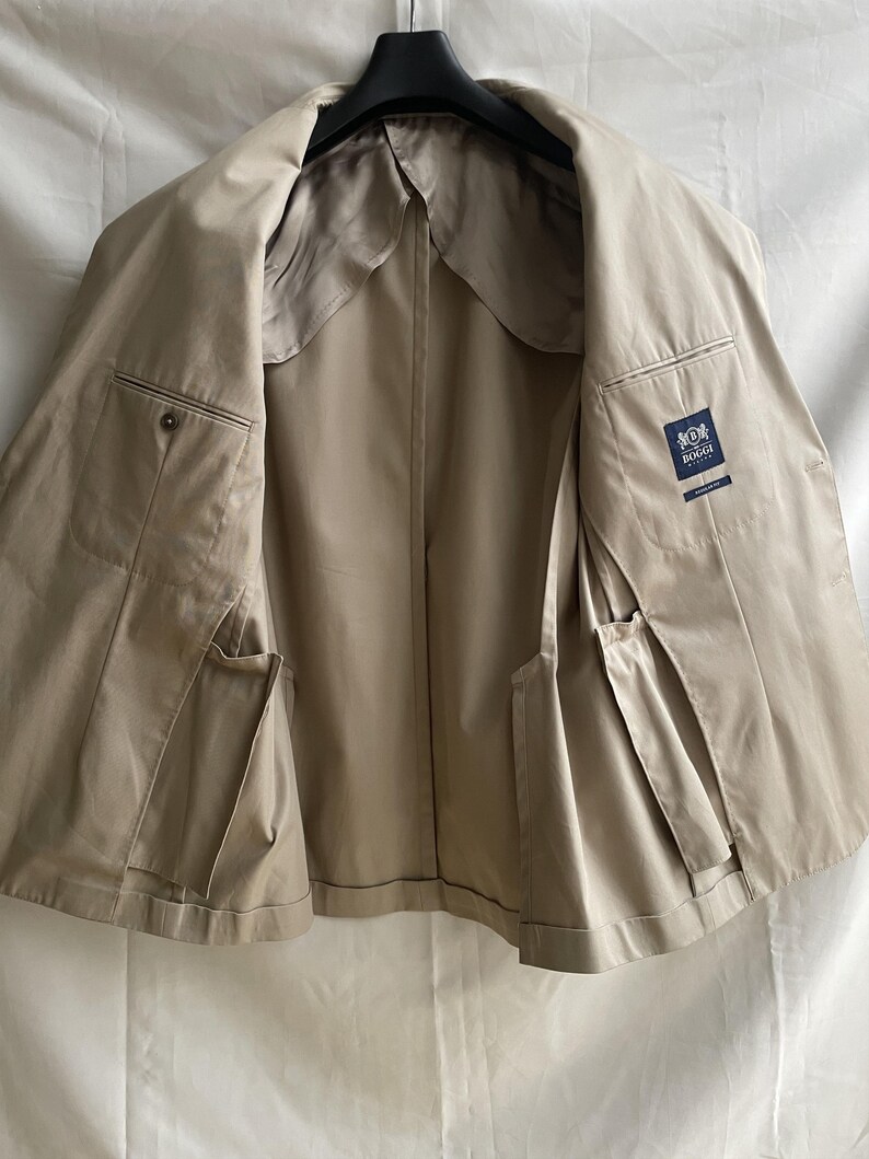 May include: A tan-colored jacket, displayed open, revealing the interior lining and pockets. The jacket is hanging on a black hanger. The interior features a satin-like lining. A label with a logo is visible on the inside.
