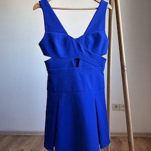 Puede incluir: Un vestido azul vibrante con escote en V y detalles recortados. El vestido est&aacute; colgado de una percha de madera, mostrando su falda plisada y corpi&ntilde;o entallado. El vestido es de color liso, perfecto para una ocasi&oacute;n especial.