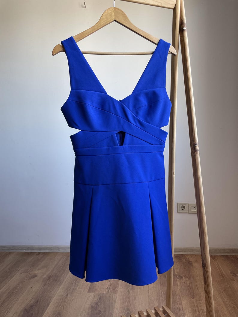 Puede incluir: Un vestido azul vibrante con escote en V y detalles recortados. El vestido est&aacute; colgado de una percha de madera, mostrando su falda plisada y corpi&ntilde;o entallado. El vestido es de color liso, perfecto para una ocasi&oacute;n especial.