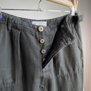 May include: Close-up of dark gray pants with a button fly and a belt loop. The pants have a high waist and a relaxed fit. The buttons are gold-colored. The pants are hanging on a wooden hanger.