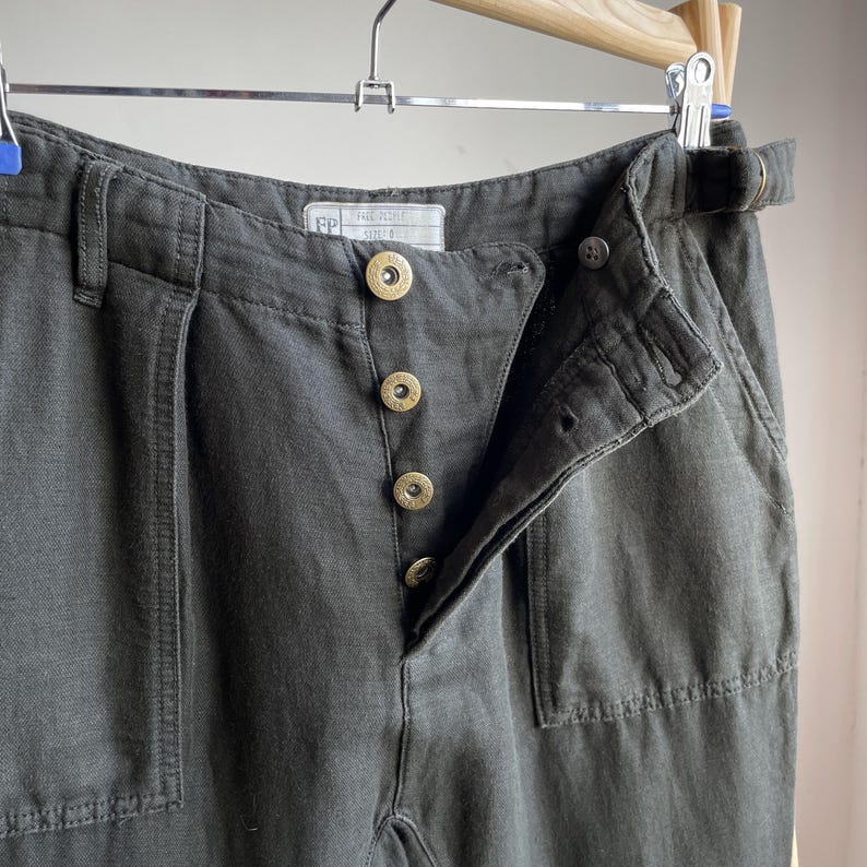 May include: Close-up of dark gray pants with a button fly and a belt loop. The pants have a high waist and a relaxed fit. The buttons are gold-colored. The pants are hanging on a wooden hanger.
