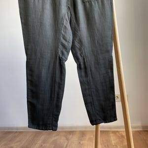 May include: Dark gray pants with a relaxed fit, featuring visible seams and a slightly faded appearance. The pants are hanging on a wooden clothing rack, showcasing their design and texture. The fabric appears to be a lightweight material.