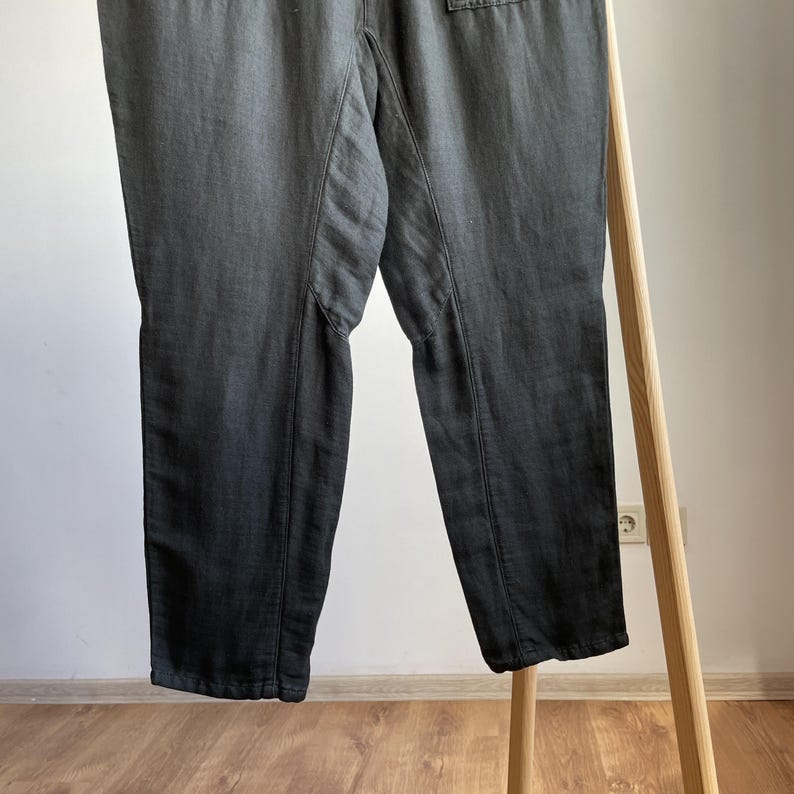 May include: Dark gray pants with a relaxed fit, featuring visible seams and a slightly faded appearance. The pants are hanging on a wooden clothing rack, showcasing their design and texture. The fabric appears to be a lightweight material.