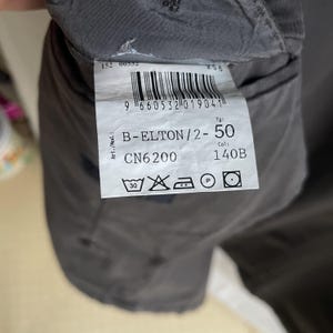 May include: Close-up of a clothing tag with the text "B-ELTON/2-50" and "CN6200". The tag also includes care symbols and a barcode. The tag is attached to a dark gray garment.
