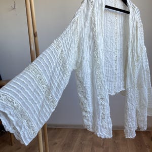 Op de afbeelding: Witte, openvallende top met lange mouwen, voorzien van een textuurstof en kanten details. De top hangt aan een zwarte hanger. De mouwen zijn wijd en zwierig, en de stof lijkt licht.
