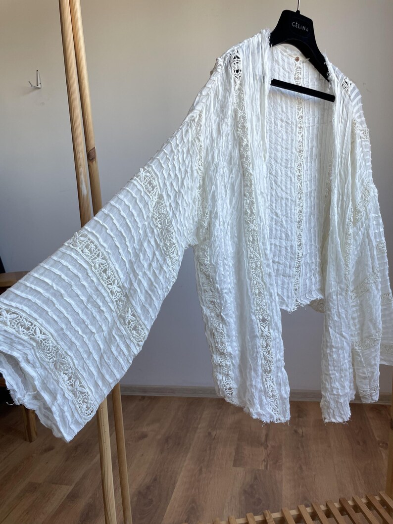 Op de afbeelding: Witte, openvallende top met lange mouwen, voorzien van een textuurstof en kanten details. De top hangt aan een zwarte hanger. De mouwen zijn wijd en zwierig, en de stof lijkt licht.