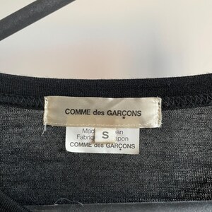 Puede incluir: Camiseta negra con una etiqueta blanca que dice "COMME des GARCONS" y "Hecho en Jap&oacute;n" con la talla "S" impresa en ella.