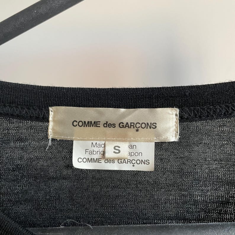 Puede incluir: Camiseta negra con una etiqueta blanca que dice "COMME des GARCONS" y "Hecho en Jap&oacute;n" con la talla "S" impresa en ella.