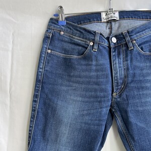 Puede incluir: Primer plano de unos vaqueros azules con un dise&ntilde;o cl&aacute;sico de cinco bolsillos. Los vaqueros est&aacute;n colgados de una percha met&aacute;lica. La marca "Acne Studios" es visible en la etiqueta.
