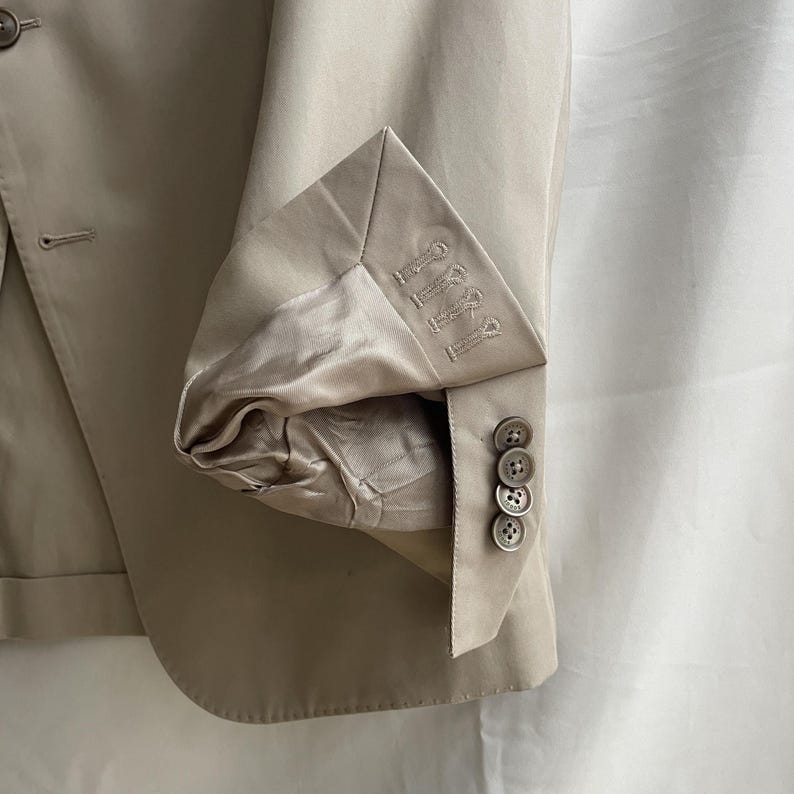 May include: A tan blazer with a folded sleeve, revealing a satin lining. The sleeve cuff features decorative stitching and four matching buttons. The blazer is made of a smooth fabric.