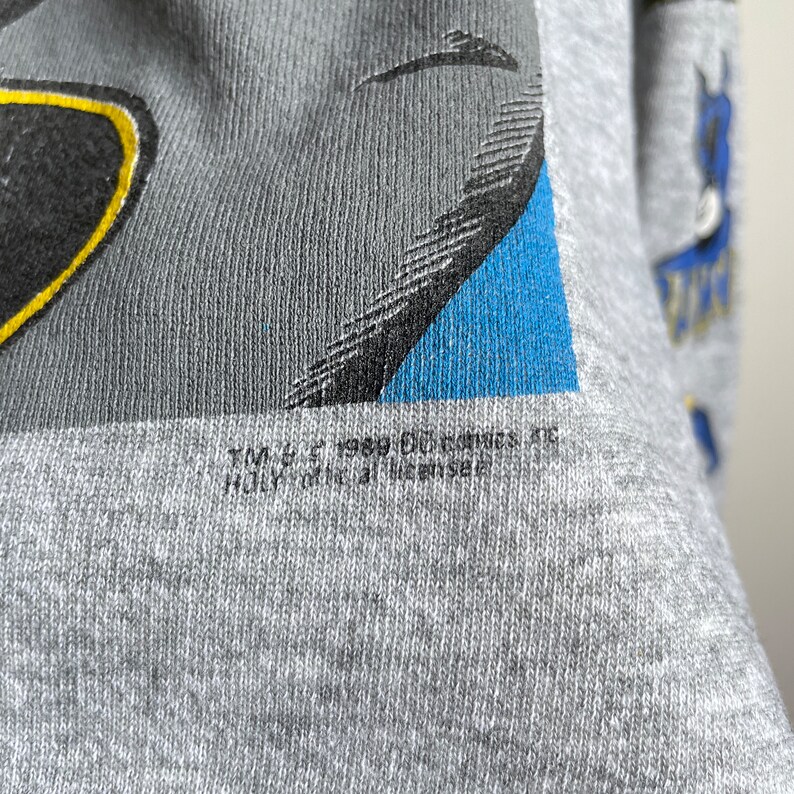May include: Grey t-shirt with a blue, black and yellow graphic of Batman. The text "TM & &copy; 1989 DC Comics. All Rights Reserved." is printed on the bottom of the shirt.