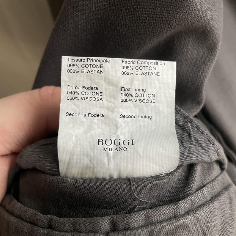 May include: Close-up of a gray garment with a fabric composition label. The label includes the brand "BOGGI MILANO" and fabric details: 98% cotton, 2% elastane for the main fabric, and 40% cotton, 60% viscose for the first lining.