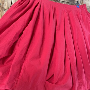 May include: A vibrant red skirt featuring a voluminous, gathered silhouette with deep vertical pleats along the waistband. The garment is suspended from a metal hanger with blue clips, showcasing a full, draped design that creates a rounded shape.