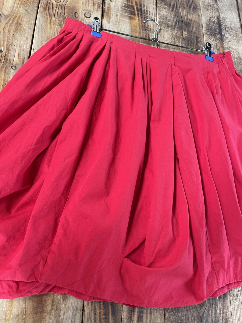 May include: A vibrant red skirt featuring a voluminous, gathered silhouette with deep vertical pleats along the waistband. The garment is suspended from a metal hanger with blue clips, showcasing a full, draped design that creates a rounded shape.