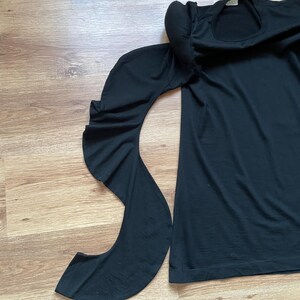 Puede incluir: Camiseta negra de manga larga con un detalle de manga con volantes. La camiseta tiene un escote redondo.