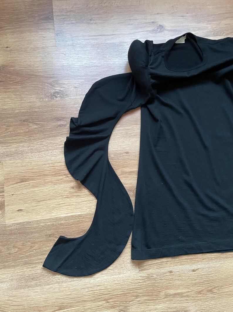 Puede incluir: Camiseta negra de manga larga con un detalle de manga con volantes. La camiseta tiene un escote redondo.