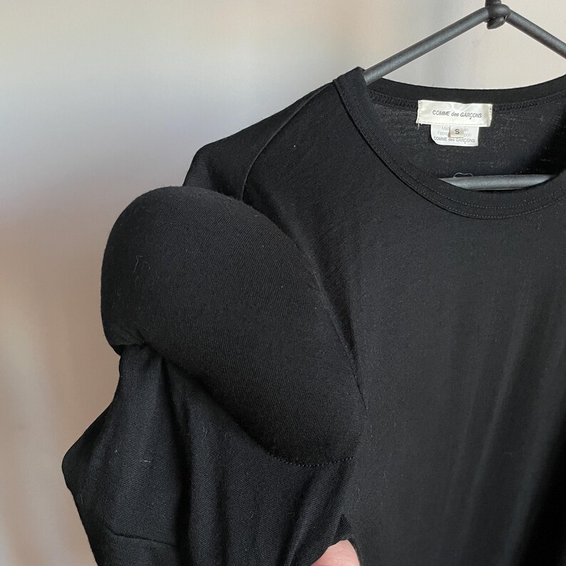 Puede incluir: Camiseta negra de manga larga con un detalle de hombro abullonado. La camiseta tiene cuello redondo y una peque&ntilde;a etiqueta blanca con el texto "COMME des GARCONS" en el lado izquierdo del escote.