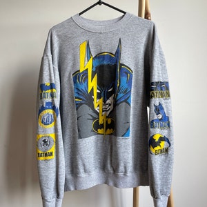 May include: Grey sweatshirt with a large graphic print of Batman on the front. The sleeves have smaller Batman graphics with the word "BATMAN" printed on them.