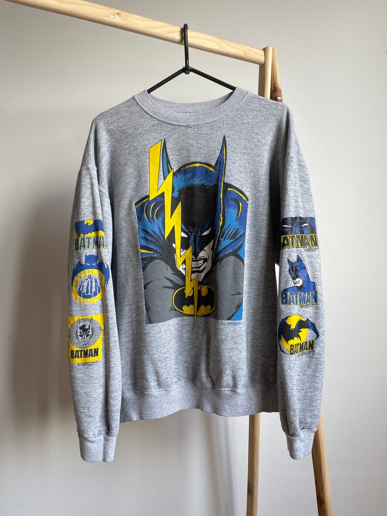 May include: Grey sweatshirt with a large graphic print of Batman on the front. The sleeves have smaller Batman graphics with the word "BATMAN" printed on them.