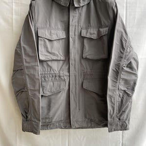 May include: A gray hooded jacket with multiple front pockets. The jacket has a full zipper closure and a drawstring hood. The jacket is made of a lightweight material and is suitable for outdoor wear. The jacket is hanging on a black hanger.