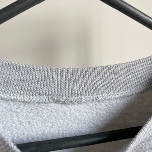 May include: Close-up of the neckline of a grey sweatshirt. The neckline is ribbed and has a sewn seam.