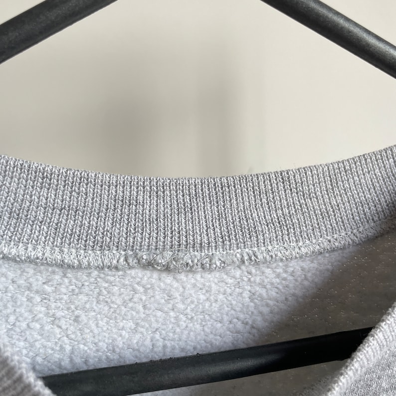 May include: Close-up of the neckline of a grey sweatshirt. The neckline is ribbed and has a sewn seam.