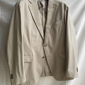 May include: A beige blazer with a notched lapel, two front pockets, and buttoned cuffs. The jacket is hanging on a black hanger against a white background. The blazer has a two-button closure.