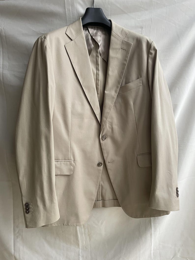 May include: A beige blazer with a notched lapel, two front pockets, and buttoned cuffs. The jacket is hanging on a black hanger against a white background. The blazer has a two-button closure.