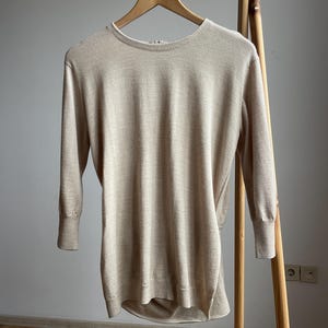 May include: A beige long-sleeved sweater with a round neckline. The sweater is hanging on a wooden hanger.
