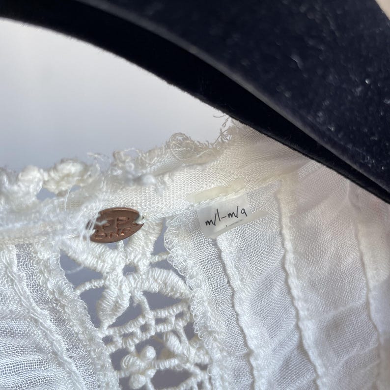 Op de afbeelding: Close-up van een witte, kanten top met een bloemenmotief. De top heeft een label met de tekst "m/l-m/q" en een klein, rond, bronzen label met de merknaam. De top hangt aan een zwarte hanger.